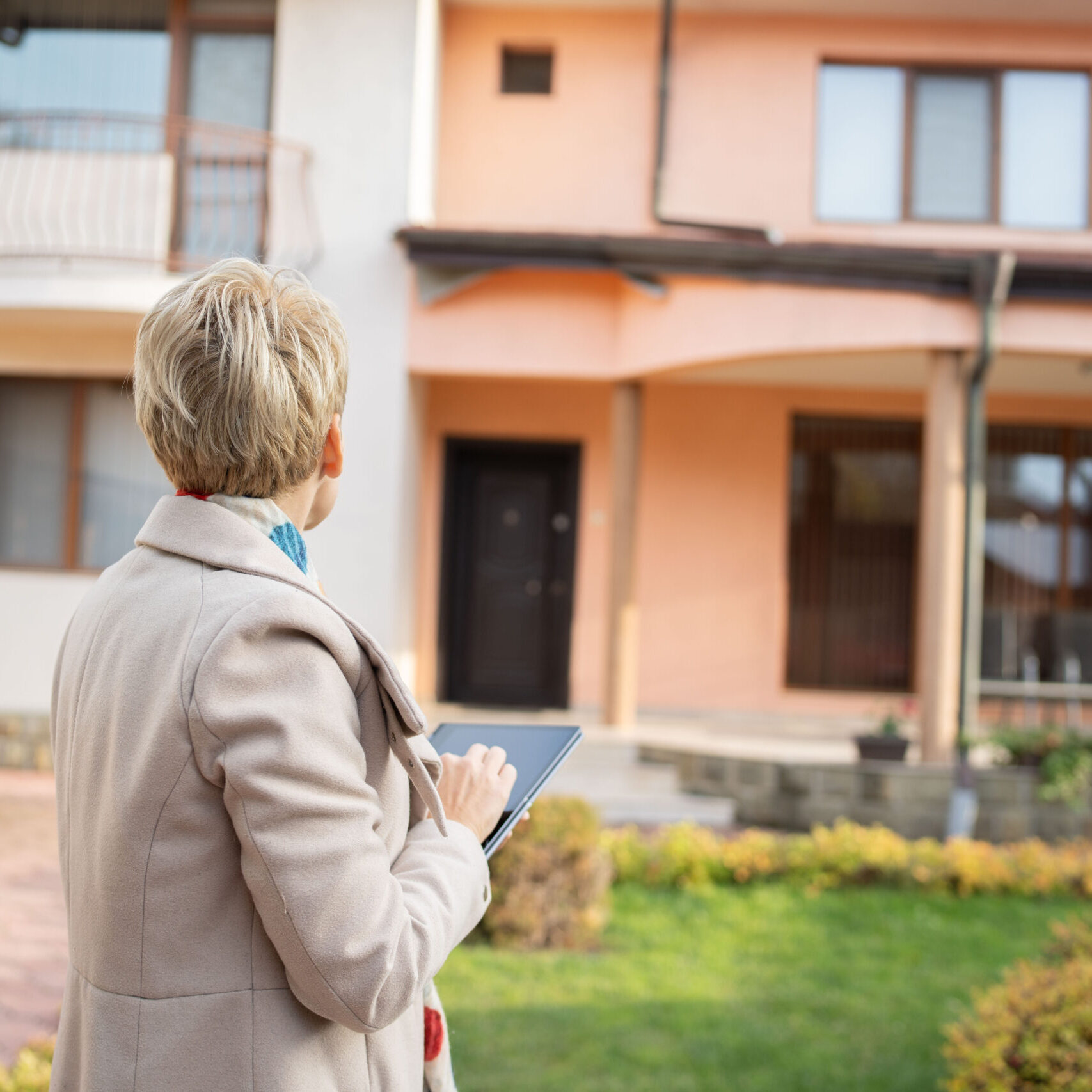 With a tranquil garden backdrop, a woman aims her tablet at the exterior of a charming home, emphasizing the role of technology in modern home exploration and decision-making.